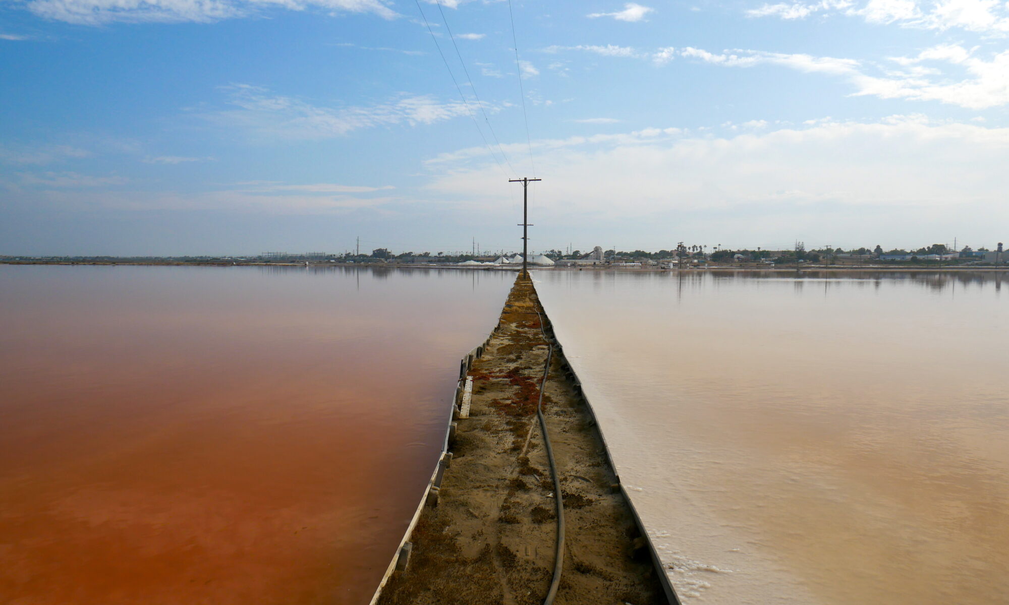 A dirt path separates two salt ponds on either side. On the left, a deep pink-red pond with still water, and on the right, a white-pink lake with still water.
