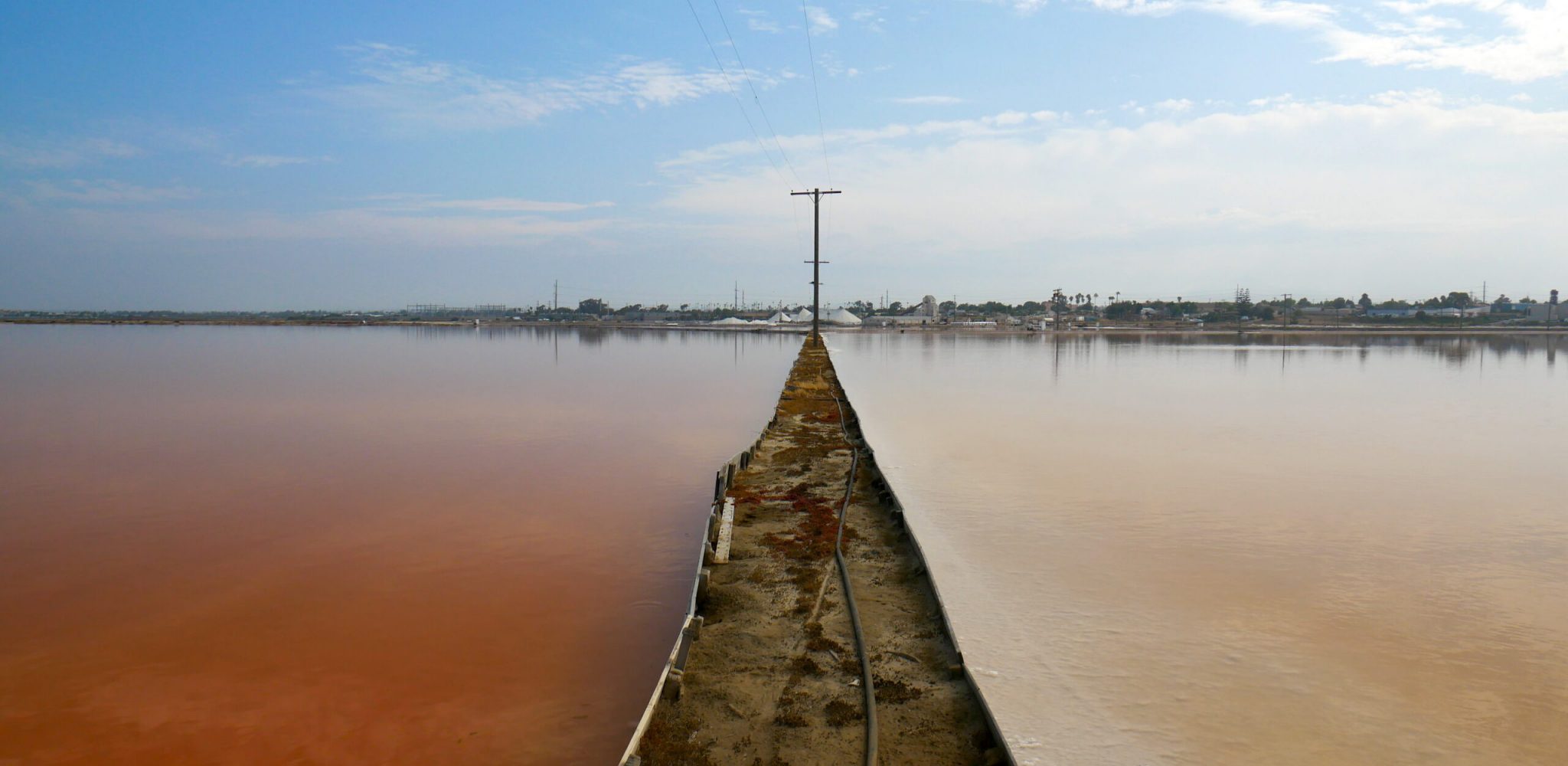 A dirt path separates two salt ponds on either side. On the left, a deep pink-red pond with still water, and on the right, a white-pink lake with still water.